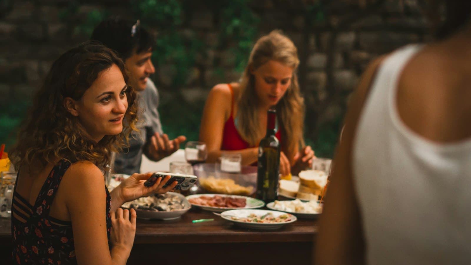 Mesa larga de madera al aire libre con personas compartiendo una comida, vasos con agua y ambiente de risas.