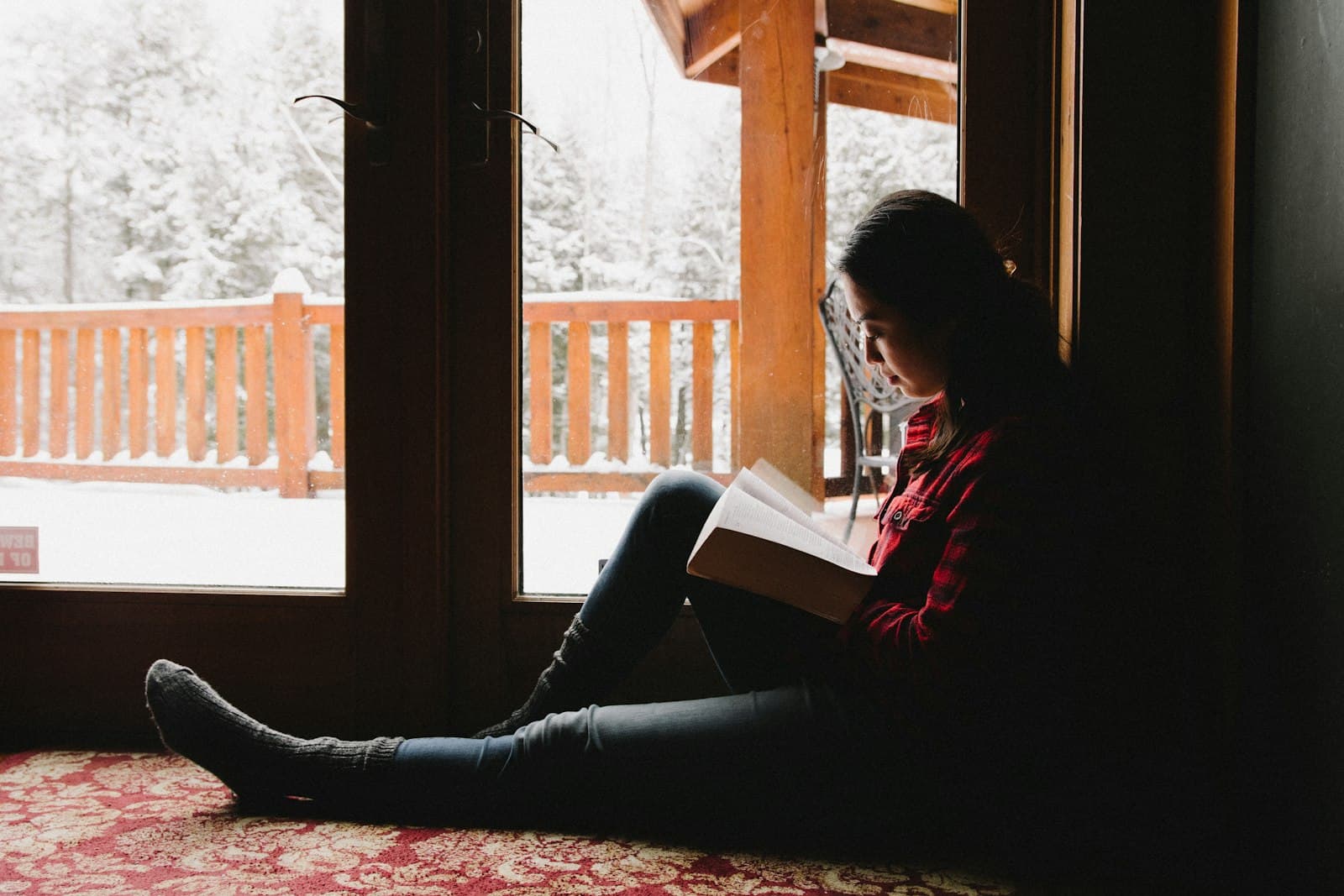Una persona leyendo un libro en una cabaña acogedora con una gran ventana que mira a la naturaleza, inmersa en la lectura.