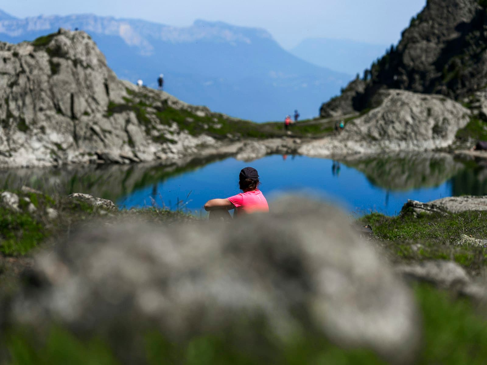 Una persona sentada tranquilamente a la orilla de un lago sereno, con montañas majestuosas al fondo.