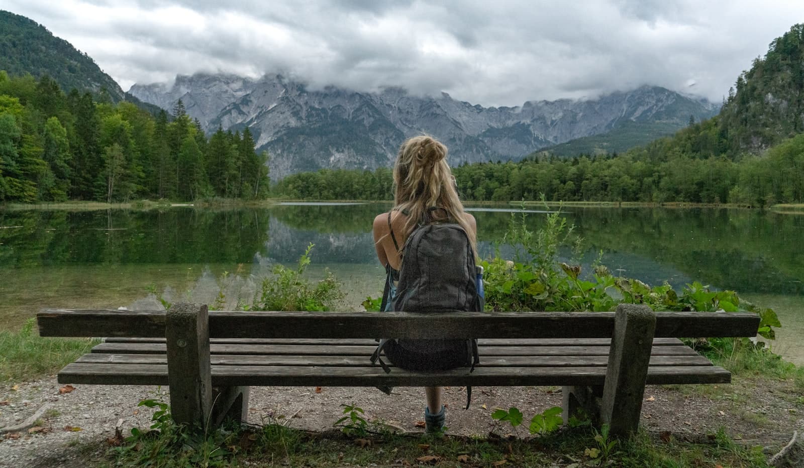 Una mujer sentada a la orilla de un lago tranquilo, contemplando las montañas distantes, en un momento de reflexión.